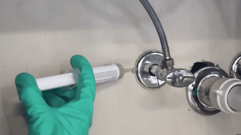 A homeowner wearing a glove and using a syringe to apply small dots of gel bait near a pipe under a sink.
