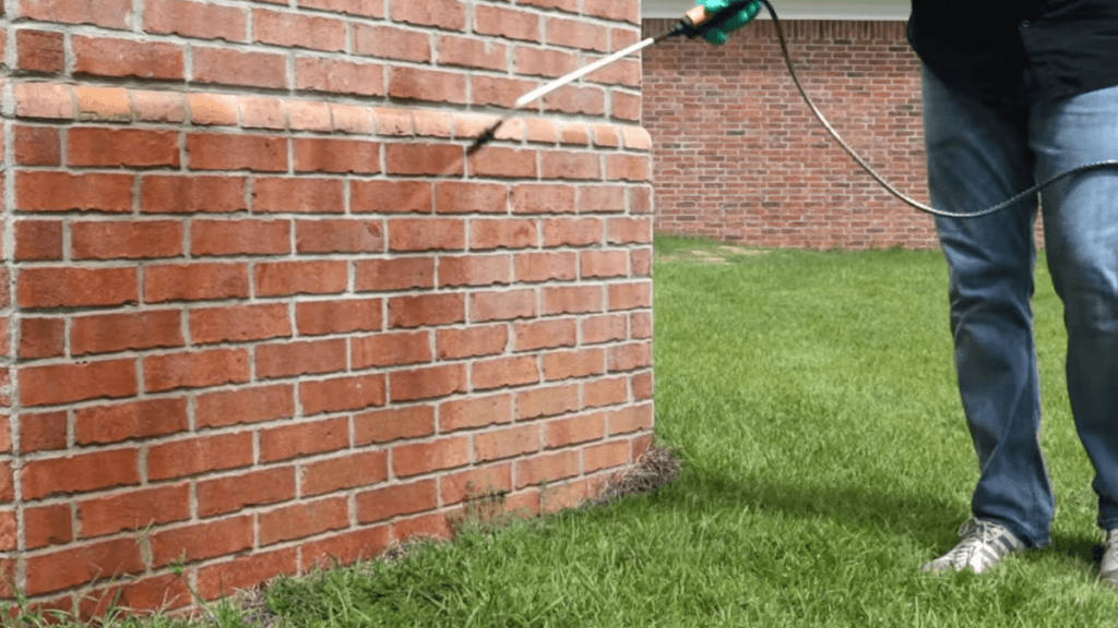 A homeowner using a hand sprayer to apply a liquid barrier treatment to the brick exterior and grass perimeter.