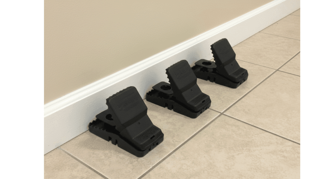 Three black T-Rex rat traps placed perpendicular to a white baseboard on a beige tile floor.