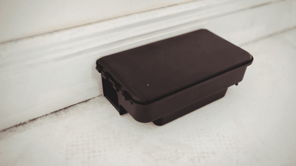 A black plastic rodent bait box placed flush against a home's exterior baseboard.