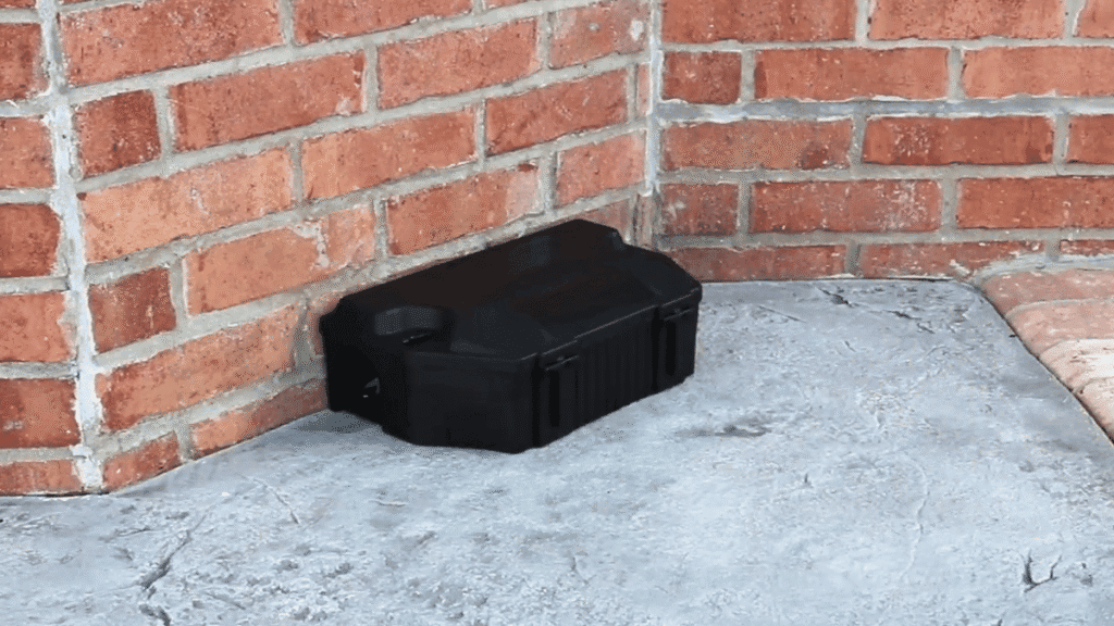 A black tamper-resistant rodent bait box placed against a brick wall on a concrete surface.