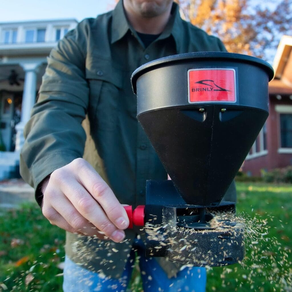 Person using a Brinly handheld crank spreader to apply seed or granules in a yard